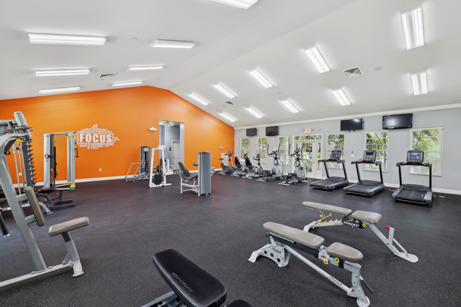 Spacious gym with treadmills, weight machines, benches, and an orange accent wall with motivational text.