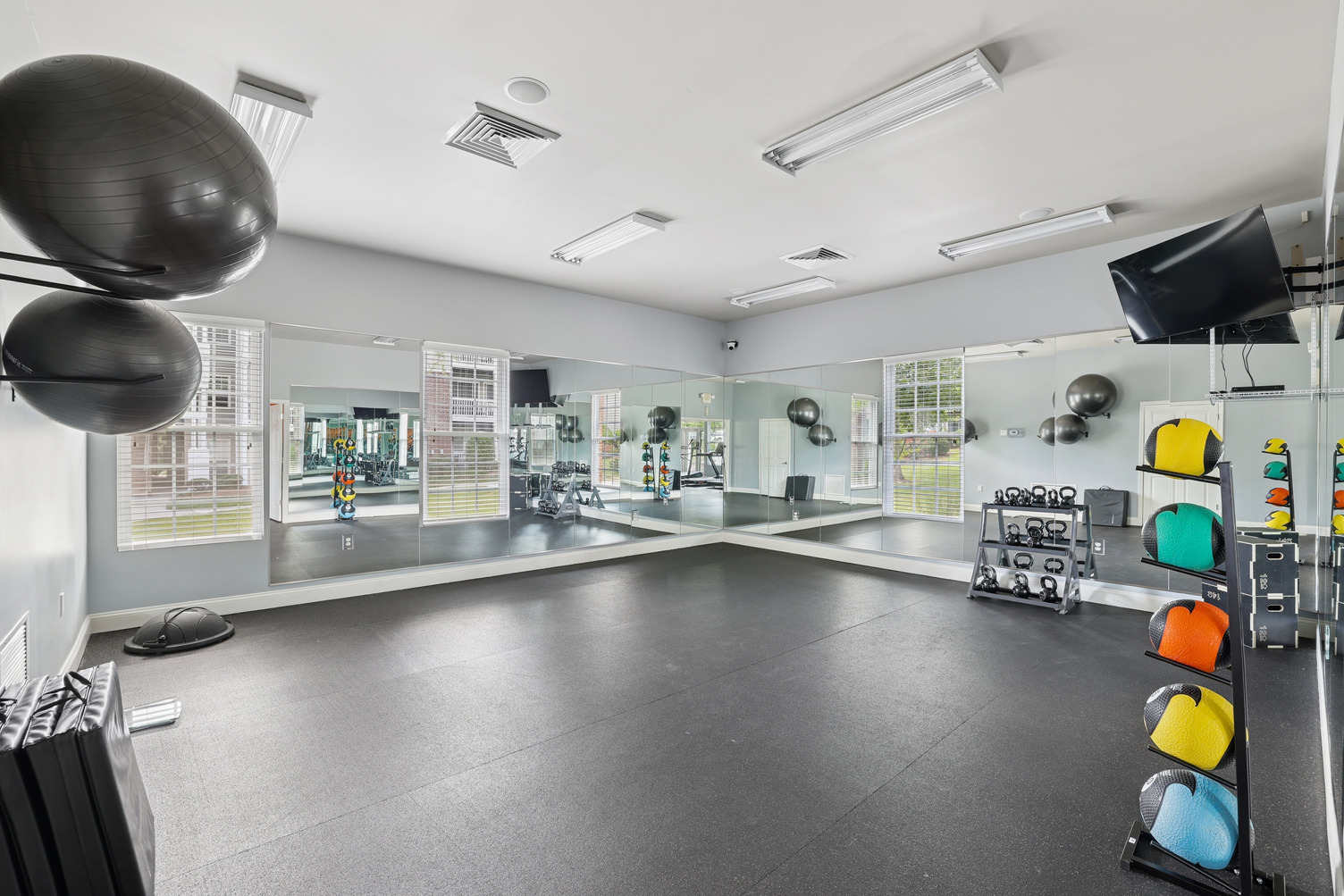 Bright gym room with large mirrors, exercise balls, weights, kettlebells, and a wall-mounted TV.