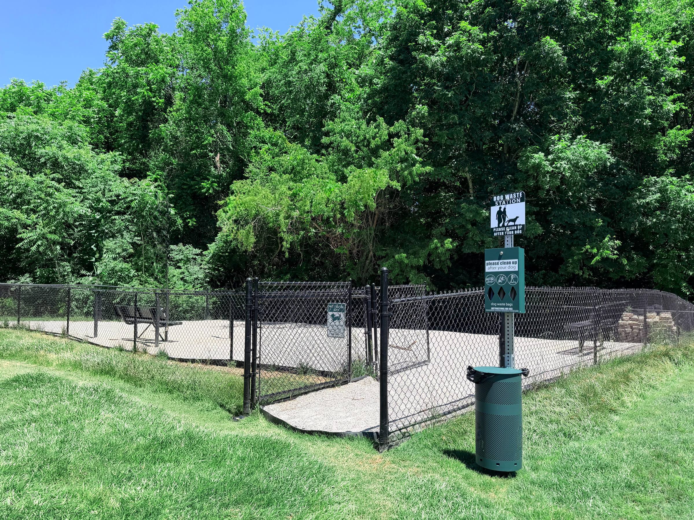 Fenced dog park with benches, a trash can, and trees in the background on a sunny day.