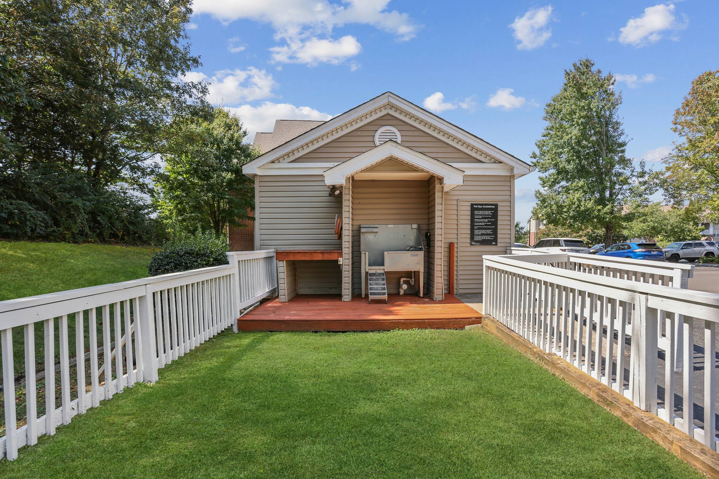 Small outdoor building with a sink and table, white railings, and a grassy area under a blue sky.