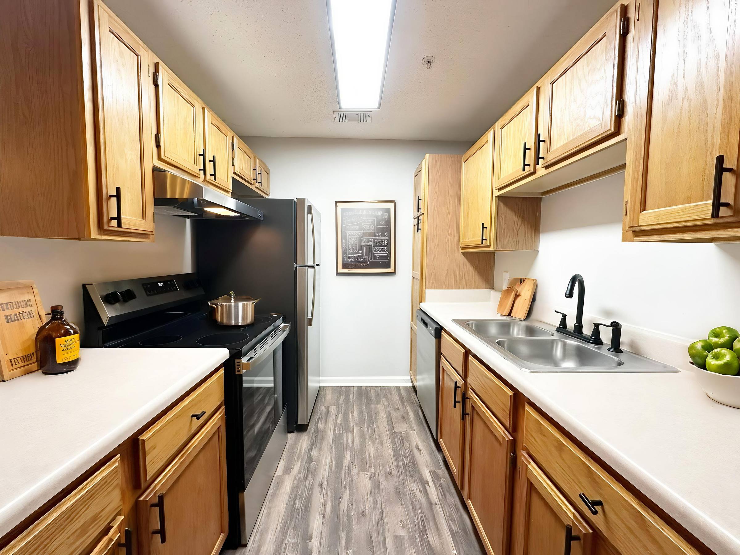 Modern galley kitchen with wood cabinets, stainless steel appliances, and a bowl of green apples on the counter.