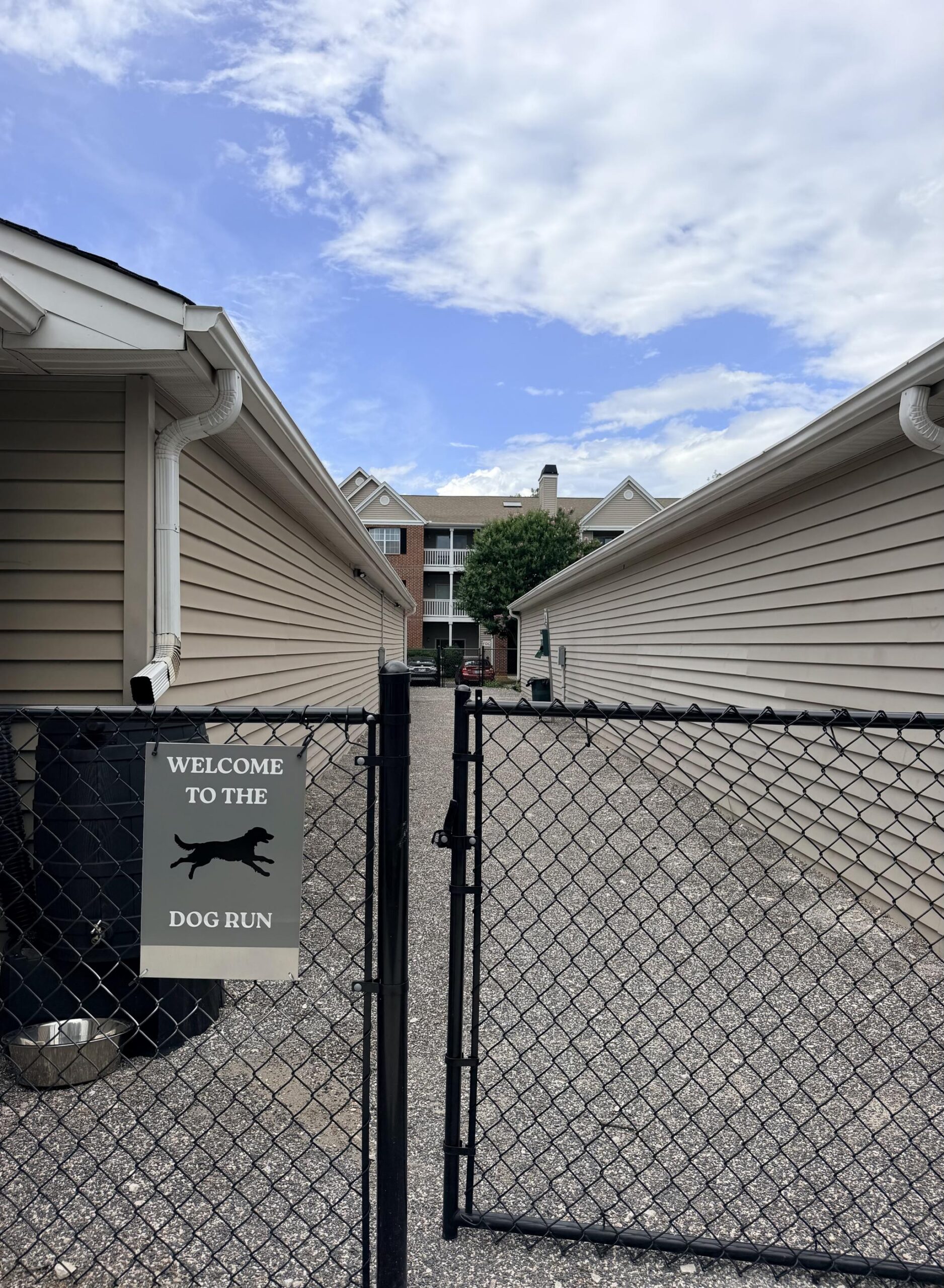 Black chain-link gate with a "Welcome to the Dog Run" sign, between two beige buildings under a blue sky.