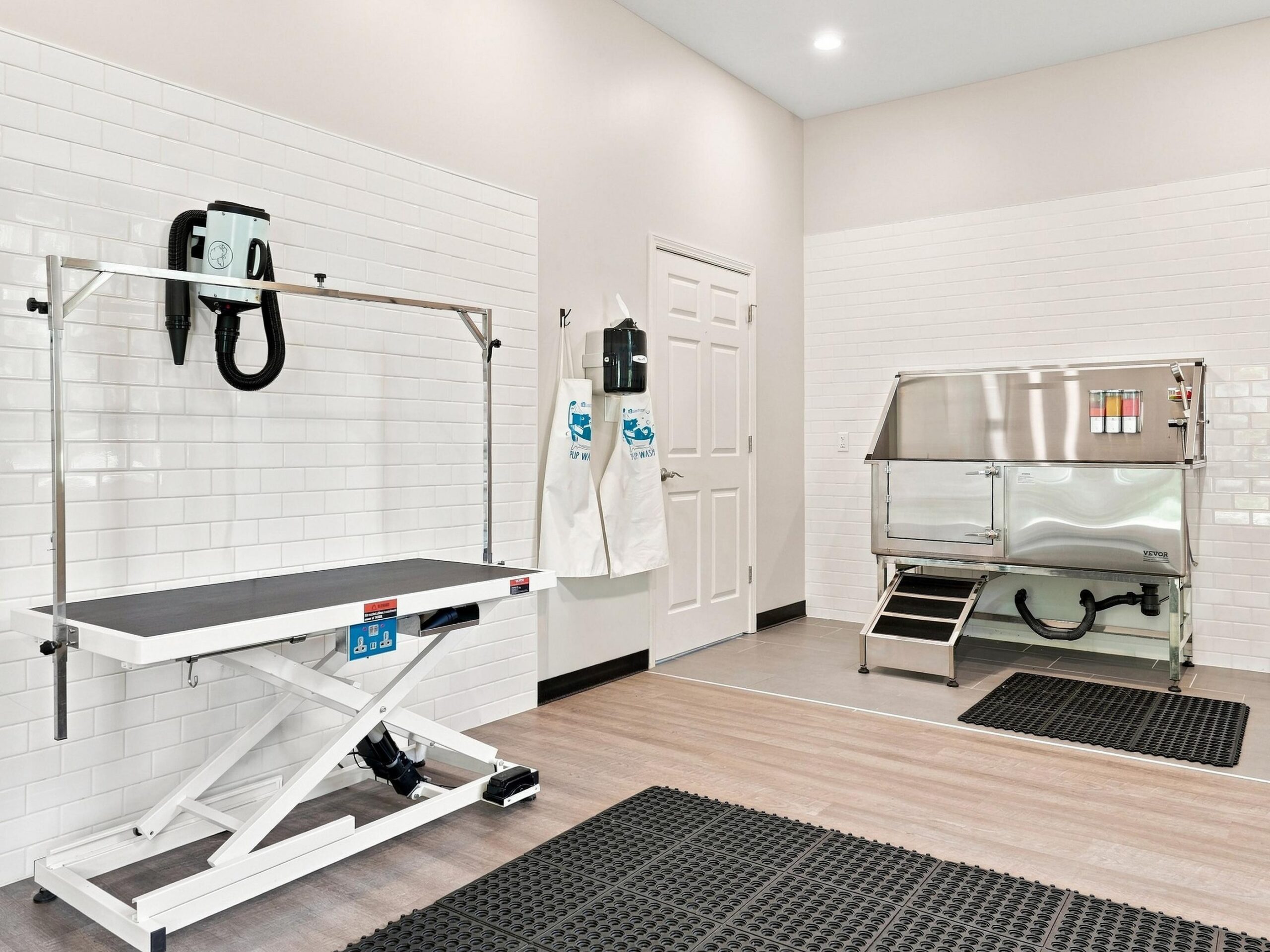 A clean pet grooming room with a grooming table, hair dryer, bath station, aprons, and mats on a wood floor.