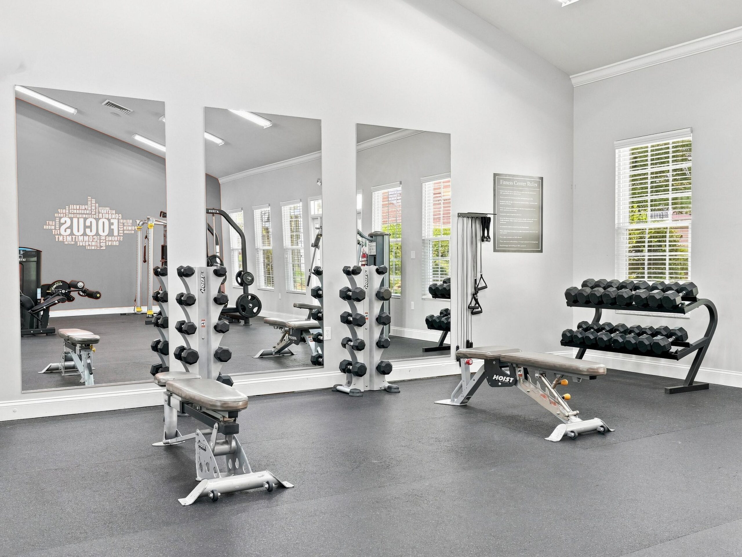 A bright gym with benches, dumbbells, mirrored walls, and windows letting in natural light.