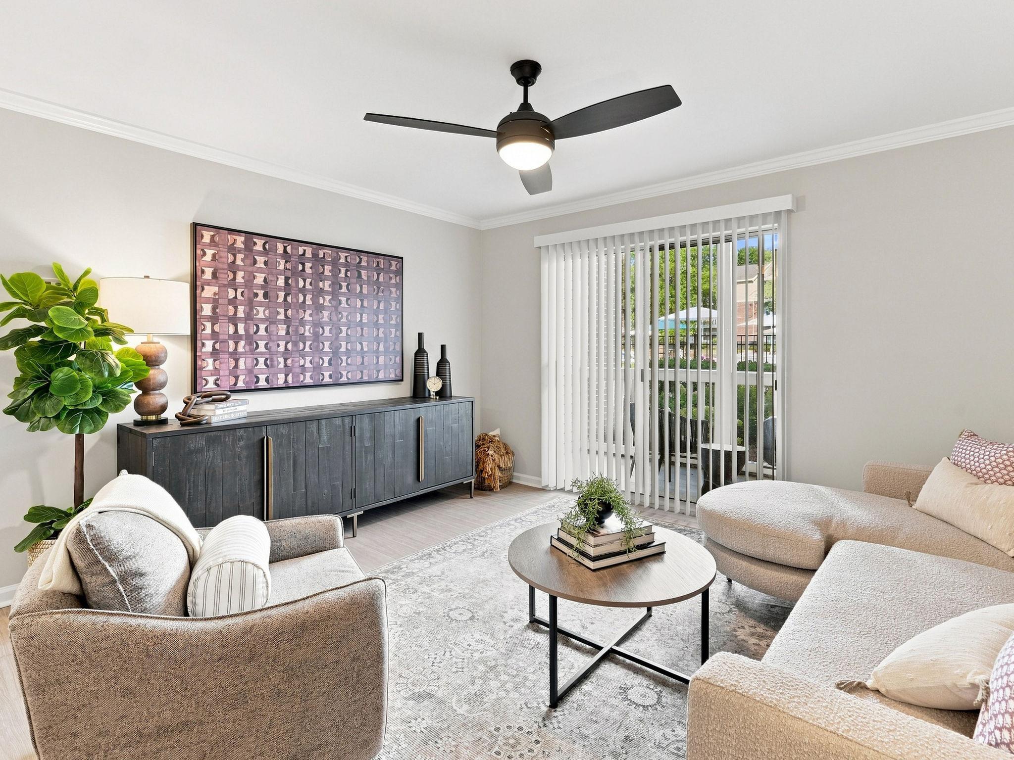 Modern living room with beige sofa, armchair, round coffee table, TV console, artwork, lamp, large plant, and sliding glass doors with blinds.