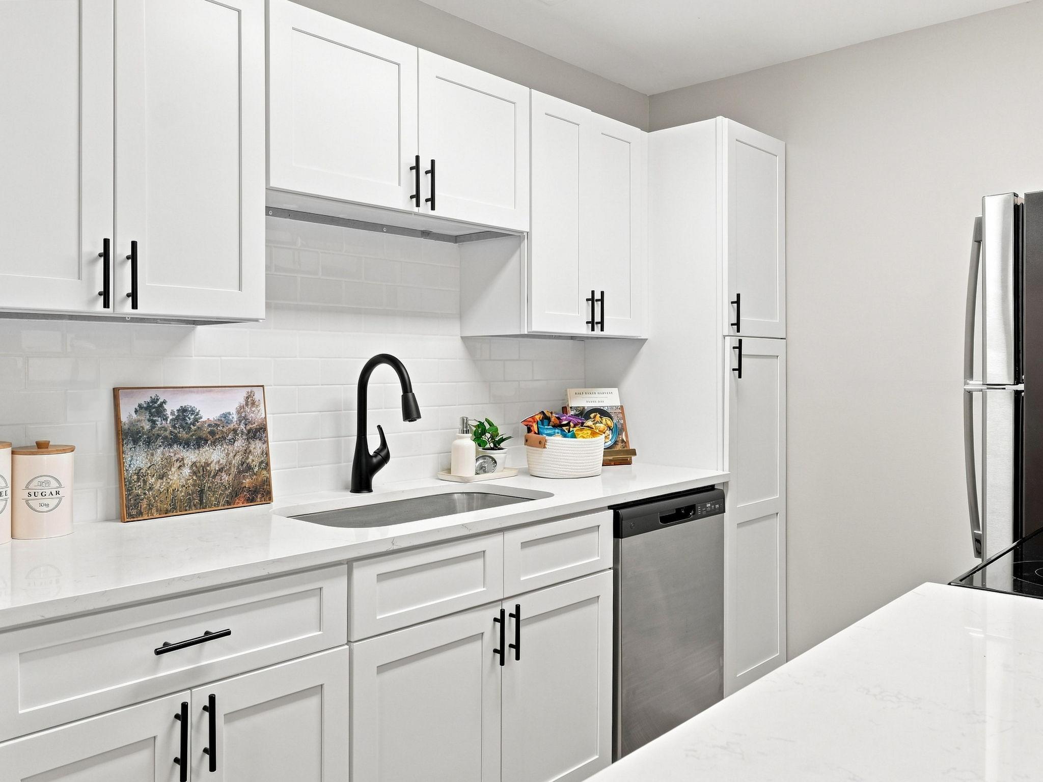 Modern white kitchen with black fixtures, stainless appliances, a bowl of fruit, framed art, and neatly arranged countertop items.