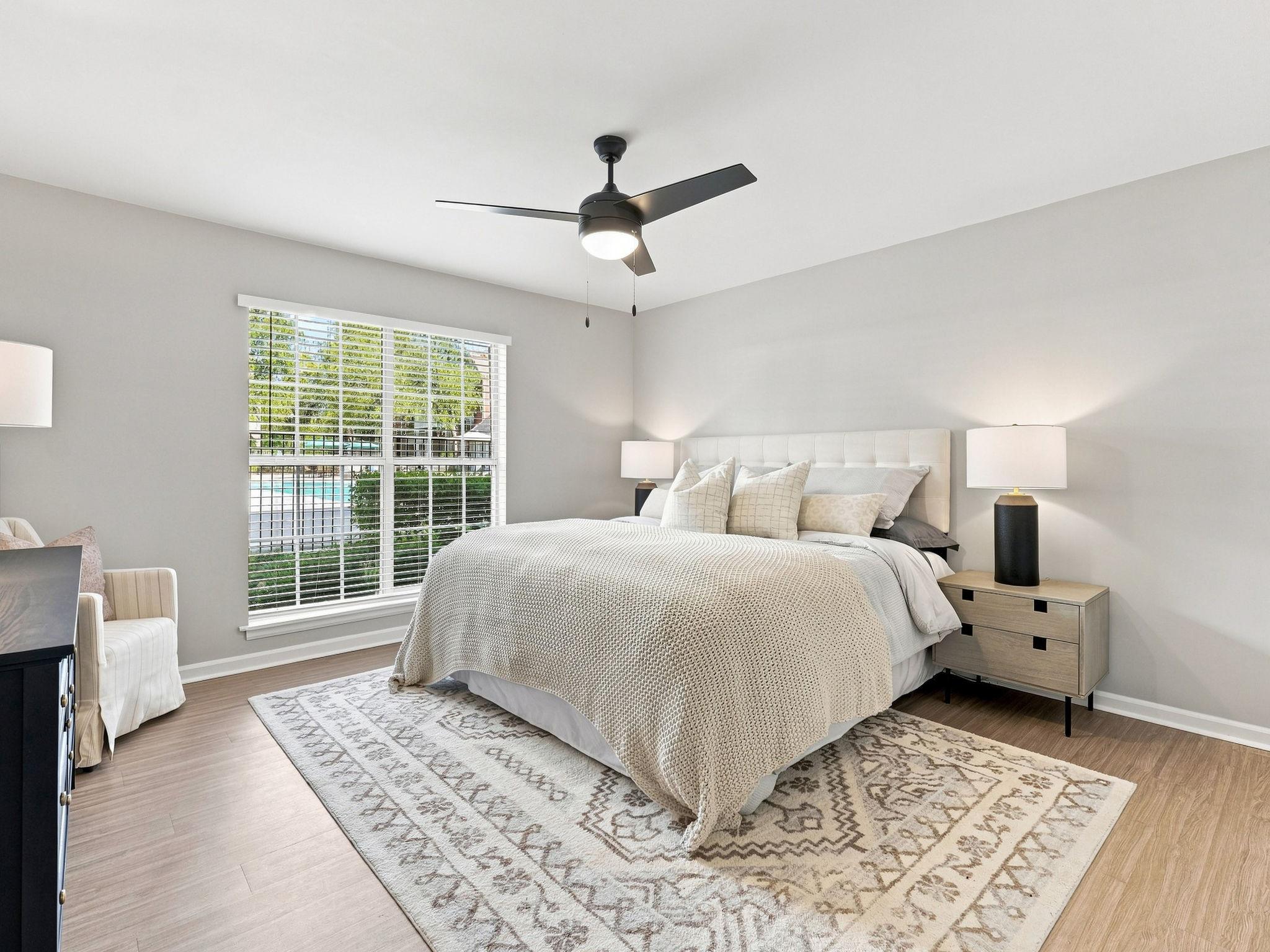 Bedroom with a large bed, two nightstands, lamps, a ceiling fan, and a window with blinds overlooking a green outdoor area.